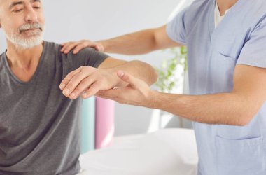 Bir rehabilitasyon terapisti tıp merkezinde yaşlı bir adamın elini destekler. Fizyoterapi ve rehabilitasyon konsepti. Bir uzman yaradan sonra hastanın işlevini yerine getirmesine yardım eder.