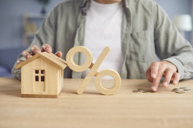 Yüzde ahşap sembol ve evin maketi arka planda bozuk paraları sayan genç bir adam. Gayrimenkul, finans, işletme, ipotek kredisi, mortgage faiz oranı kavramı