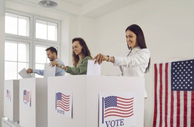 Amerikan vatandaşlarının portresi oy sandığında sırada duruyor ve oy pusulalarını oy kabinine koyuyor. Mutlu insanlar seçim günü oy kullanma merkezinde ABD bayraklarıyla oy veriyorlar..