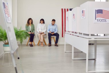 Oy verme merkezinde Amerikan bayrağıyla süslenmiş bir sıra beyaz oy kullanma kabiniyle oy kullanma merkezi. Amerikan seçmenleri sandık merkezinde oturuyorlar. Demokrasi ve seçim günü konsepti.