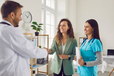 Tıbbi muayeneden sonra gülümseyen erkek doktor, genç ve mutlu hasta bir kadınla el sıkışıyor. İlaç ve sağlık hizmetleri kavramı.