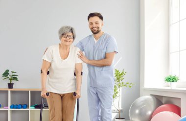 Klinikteki bir doktor yaşlı bir kadının koltuk değnekleriyle yürümesine yardım eder, yakın plan. Yaralanmadan sonra iyileşme süreci. Klinikte profesyonel tıbbi bakım, rehabilitasyon süresince destek