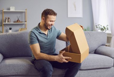 Mutlu adam evdeki oturma odasındaki kanepede oturuyor, online alışveriş siparişinden bir kutu, paket ya da paketi hevesle açıyor. İçindekilerden memnun, teslimatın sevincinden zevk alıyor..
