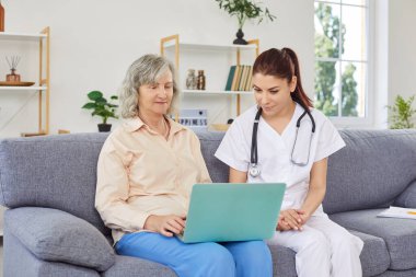 Dost canlısı genç bir hemşire ya da doktor yaşlı bir kadına evdeki kanepede otururken dizüstü bilgisayar kullanmasında yardım ediyor. Emekliler için tıp, sağlık ve evde bakım kavramı.