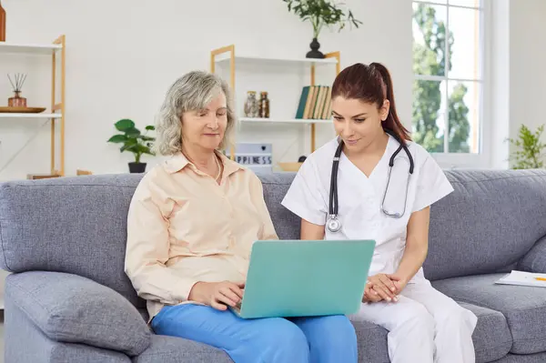 stock image Friendly young woman nurse or doctor helping senior elderly female person in using laptop sitting on sofa at home. The concept of medicine, health and home care for retirement people.