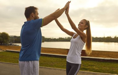 Erkek koç ya da antrenör, şehir parkında spor yaparken spor eğitimi alan mutlu stajyer bir kadına beşlik çakıyor. Zindelik yolculuğunda beraberlik teşvik ve destek.