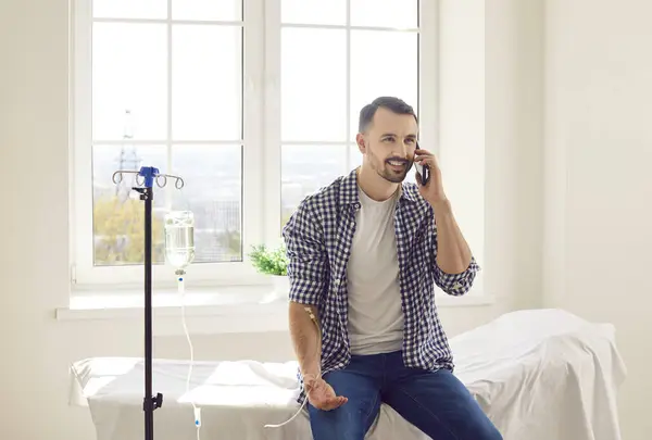 stock image Young man sitting on the couch in clinic talking by mobile phone while receiving IV drip infusion and vitamin therapy. Smiling male person receiving injection therapy in medical office.