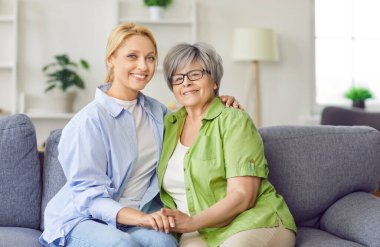 Nazik, sevgi dolu, olgun yaşlı bir anne, yetişkin bir kız, kanepeye yakın oturan, şefkatli genç bir kadın, yaşlı bir anneyi kucaklayan, mutlu uluslararası kadınlar gününde, evde anneler günü tatilinde eğlenen.