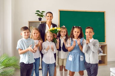 İlkokul çocuklarının ve sınıf öğretmenlerinin şenlikli portresi. Çiçekleri olan mutlu bir kadın ve öğrencileri kara tahta arkasında kameraya gülümsüyor..