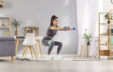 Genç sportif bir kadın iki bacaklı derin bir sabah egzersizi yapıyor. Oturma odasında dambıllarla duruyor. Fit kız yağ kalorileri yakıyor. 