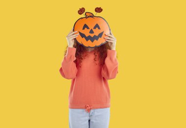 Cadılar Bayramı 'nda komik kağıt balkabağının arkasında yüzünü saklayan neşeli, tanınmayan bir kadın. Uzun kırmızı kıvırcık saçlı, rahat kot pantolon giyen ve turuncu arka planda izole edilmiş sweatshirt giyen genç bir kadın..