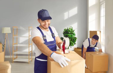 Genç neşeli işçi, yeni bir daireye taşınmayı planlayan yapışkan bantlı karton kutu paketliyor. Taşımacılık işlerinde çalışan erkek bir ekip. Yerleştirme ve emlak konsepti.