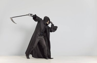 Ölüm meleği kostümlü neşeli komik adam siyah pelerinli ve iskelet maskeli salıncak tırpanı gri arka planda garip bir poz veriyor. Cadılar Bayramı 'nda acımasız bir adam eğleniyor ve korkutuyor..