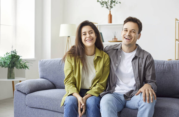 Портрет счастливой семьи молодой пары, сидящей вместе на диване в гостиной. Они радуются и улыбаются перед камерой, показывая любовь и радость в домашней обстановке.