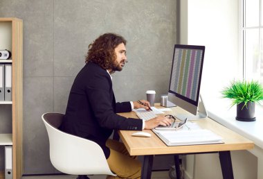 Side view of a young stylish bearded business man working on a laptop with tables and charts at the office or at home and making calculations. Business accounting, data analysis and finance concept. clipart