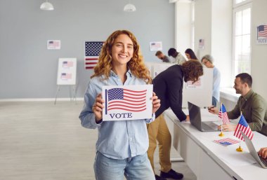 Gururlu Amerikalı seçmenler ABD 'deki başkanlık seçimleri sırasında oy vermeye geliyor. Amerikan bayrağıyla VOTE posteri taşıyan mutlu, güzel, gülümseyen genç bir kadının portresi.