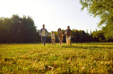 Mutlu aile koşuları ve yaz parkında eğlenmeler. Doğanın güneşli günlerinin tadını çıkarıyorlar. Anne, baba ve neşeli iki çocukları gün batımında yeşil çimlerde yürüyorlar..