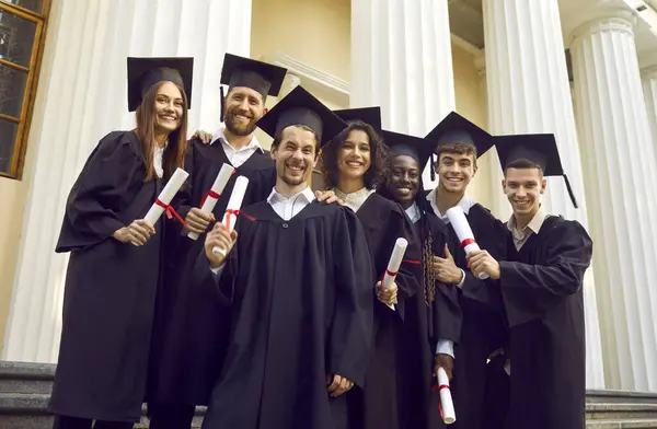 stock image Portrait of a smiling happy diverse multiracial international graduates students standing in a university graduate gown and holding diploma outdoor. Education and graduation concept.