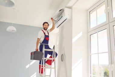 Profesyonel elektrikçi ya da teknisyen portresi merdivenin üzerinde duran, kameraya bakan ve gülümseyen, bakımlı, temizleyen, tamir eden ya da modern klima takan.