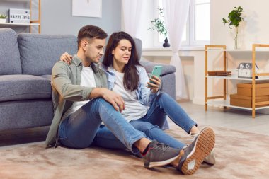 Genç pozitif aile çifti boş zamanlarını evde geçirir, yerde dinlenir, internet eğlencesi için cep telefonu kullanır. Beraberlik ve rahatlama, çevrimiçi aktivitelerin keyfi.