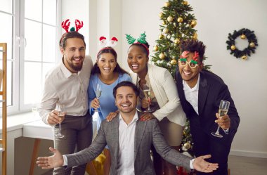 Yeni yıl partisinde birlikte poz veren neşeli, çok kültürlü genç insanların portresi. Noel aksesuarları takan ve ellerinde şampanya bardakları olan erkekler ve kadınlar ev partisinde kameraya gülümsüyorlar..