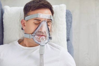 Yukarıdan yakından, yüzünde tıbbi oksijen maskesi olan hasta bir adam evdeki kanepede yumuşak bir yastığın üzerinde yatıyor. Anea bozukluğu olan yetişkin bir erkek uyurken CPAP maskesi takıyor. Hastalık tedavisi kavramı