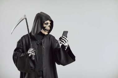 Korkunç Azrail, tırpanlı ve iskelet maskeli gri arka planda bir cep telefonu kullanıyor. Bu ürkütücü görüntü Azrail 'in ölümcül varlığını internet elementleriyle birleştiriyor..