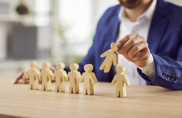 Businessman wearing suit pulling man wooden toy figure from the crowd of employees standing in a row sitting at desk in office. Business, human resource management, recruitment and leadership concept