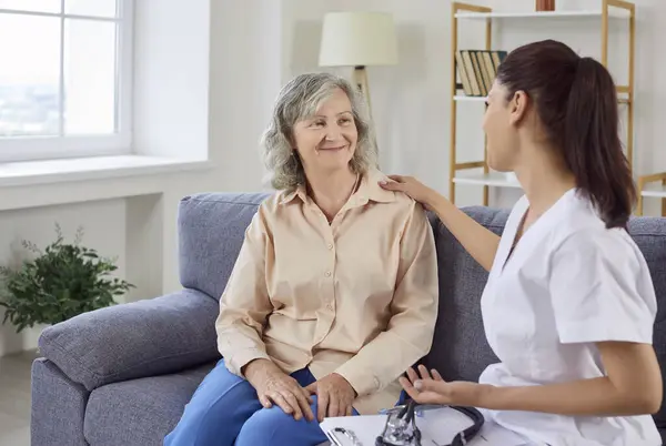 stock image Portrait of sick senior woman sitting at on sofa at home with a young female nurse supporting her. Female elderly patient listening a doctor visiting her during medical exam. Health care concept.