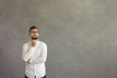 Beyaz gömlekli, gözlüklü, düşünceli, tereddütlü ve şüpheci yakışıklı bir genç. Zeki yüz ifadesi olan düşünceli adam gri beton kopya alanı arka planında dikiliyor.