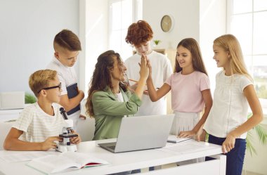 Gülümseyen öğretmen bir grup öğrenciyle bir okul sınıfında ders veriyor ve onlara bir beşlik çakıyor. Öğrenciler, öğrenim ortamında birlik ve takım çalışması sergileyerek birbirlerine bağlı ve özenli davrandılar..