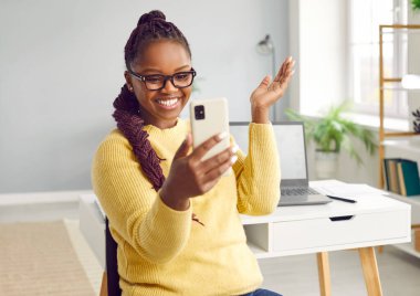 Mutlu gülümseyen Afrikalı-Amerikalı güzel kız cep telefonuna bakıyor, oturma odasında dizüstü bilgisayarla masanın arkasında duruyor. Cep telefonundan harika haberler alan mutlu bir kızın portresi.