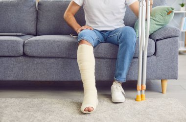 Kırık bacaklı bir adam oturma odasındaki kanepede oturur, yakın plan. Kavram yaralanması, kemik kırığı, tedavi ve evde rehabilitasyon, kaza. Koltuk değneği kullanımı, tıbbi bakım, travma bilimi.