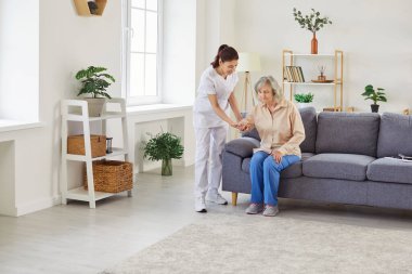 Genç bir hemşirenin portresi yaşlı bayan hastaya evdeki kanepeden kalkması için yardım ediyor. Fizyoterapist yaşlı kadına rehabilitasyonda yardım ediyor. Sağlık çalışanı yaşlı bir insanın yürümesine yardım ediyor.