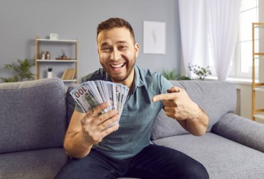 Zengin adam finansal başarısını kutluyor, evdeki bir kanepede oturuyor ve elinde yığınla nakit para tutuyor. Mutluluk ve memnuniyet, kazanma ve zenginliğin tadını çıkarma hissini somutlaştırıyor..