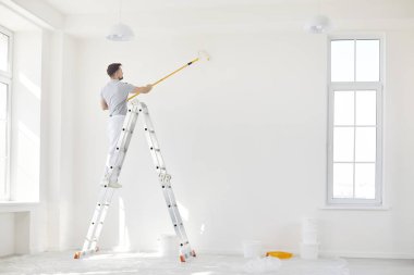 Boyacı olarak çalışan bir adam, evin tadilatı sırasında bir duvarı boyamak için paten fırçası kullanıyor. Bu yapı, ev geliştirme projelerinde yer alan ayrıntılı ve temel çalışmaları vurguluyor.