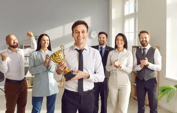 stock image Smiling Caucasian businessman holds a champion cup, surrounded by colleagues who are applauding him. Celebration, team support and recognition of success and achievement in the office.