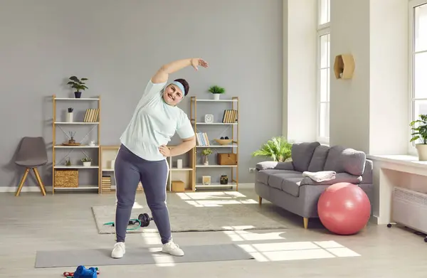 stock image Overweight, chubby woman doing side bends at home. Plus size woman in sportswear and headband doing fitness workout. Weight loss workout, domestic training, healthy lifestyle