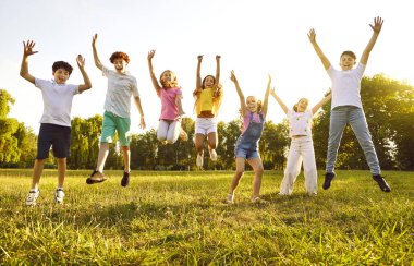 Bir grup güler yüzlü çocuk parkta yeşil çimlerin üzerinde sırayla zıplıyorlar. Mutlu çocuklar güneşli bir yaz gününde kampta birlikte eğleniyorlar..