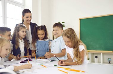 Dost canlısı gülümseyen bayan öğretmen ilkokuldaki çocuklara eğlenceli bir ders vermekten mutluluk duyar. Kalemleri ve kitapları olan neşeli okul çocukları bir masada durup öğretmenleriyle konuşuyorlar..