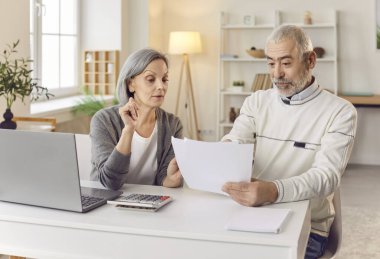 Yaşlı çift evdeki bir masada birlikte oturur ve çalışır, faturaları, belgeleri, ödemeleri gözden geçirir ve mali durumlarını yönetir. Finansal meseleleri halletmek için ortak bir çaba içindeler..