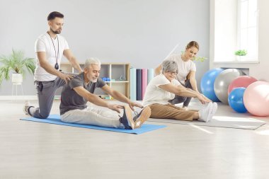 Rehabilitasyon merkezinde yoga minderinde oturan spor antrenörlerinin yardımıyla esneme sporları yapan olgun insanlar. Son sınıf emekli erkek ve kadın spor salonunda sağlık için egzersiz yapıyorlar..