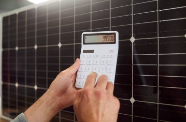 Arka planda hesap makinesi ve güneş panelleri tutan insan ellerine yakın çekim. Ekonomi ve güneş enerjisi kullanarak tasarruf. Enerji tasarrufu, yenilenebilir enerji, harcamaların azaltılması ve ucuz güneş enerjisi