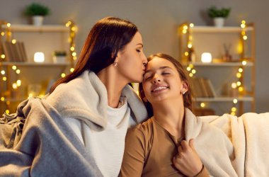 Anne koşulsuz sevgisini ifade ediyor, kızını alnından öpüyor. Mutlu anne ve çocuk, keyifli, uyumlu ve hassas duygularla dolu kaliteli bir haftasonu boyunca oturma odasında Xmas ışıklarıyla ekose kumaşlarla sarılıyorlar.