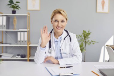 Dost canlısı gülümseyen sarışın doktor kameraya bakıyor. İnternetten arıyor ya da konferans salonunda iş arkadaşını ya da hastasını selamlıyor. Teletıp ve hasta danışmanlığı online konsept.