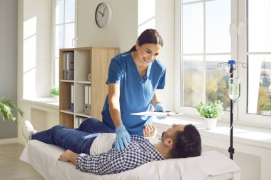 Klinikte erkek hastayla ilgilenen dost canlısı bayan hemşire. Önlüklü gülümseyen genç bir kadın elinde bir pano ve yanında duran bir adamla vitamin terapisi görüyor.