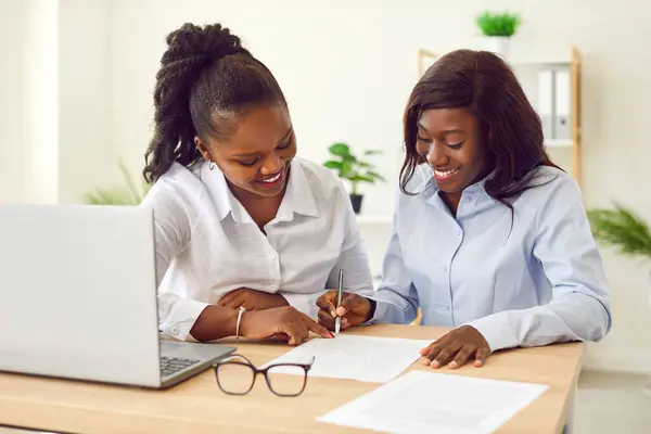 Stock image Portrait of two african american female coworkers and company employees sitting at the desk reaching agreement on meeting. Confident young business women make a deal signing a contract at the office.