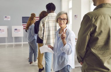 Mutlu Amerikalı seçmenler seçim günü oy kullanma merkezinde kayıt olurken zafer jesti yapıyorlar. Oy verme merkezinde kuyrukta bekleyen gülümseyen kadın kameraya neşeli bakıyor..