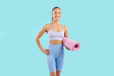 Antrenmana hazır, elinde paspas tutan mutlu ve aktif bir kadın sporcunun portresi. Mavi arka planda, spor malzemeleriyle fitness ve jimnastik coşkusunu somutlaştırıyor..