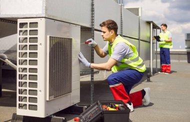 Üniformalı genç teknisyen binanın çatısında duran tornavidalı bir tornavida kullanarak klima kurup tamir ediyor. Tamirci havalandırma sistemini tamir ediyor..